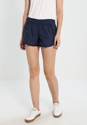 Femme portant un short de sport bleu marine, un haut sans manches blanc et des baskets blanches à semelles naturelles, debout devant un fond uni.