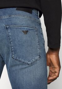 Blå denimjeans med en närbild av bakfickan som har en svart logotypaccent. Tyget har en slät textur och klassisk passform.