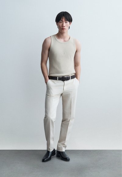 Νεαρός άνδρας στέκεται με τα χέρια στις τσέπες φορώντας μπεζ αμάνικο τοπ, ανοιχτόχρωμο παντελόνι, μαύρη ζώνη και μαύρες μπότες πάνω σε μονόχρωμο φόντο.