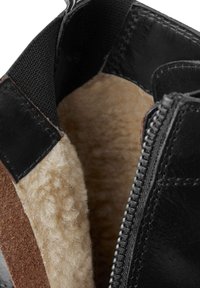 Interior de bota de cuero negra con forro de felpa marrón, que presenta una cremallera en un lado y un acento de ante marrón en la abertura.