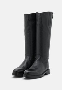 Une paire de bottes noires en cuir hautes avec des bouts ronds et des semelles plates, se tenant debout sur un fond blanc.