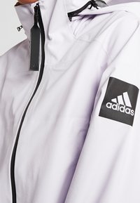 Acercamiento de una chaqueta blanca de Adidas con una cremallera negra y un parche del logo de Adidas en la manga izquierda.
