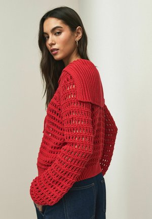 Hosszú sötét hajú nő, aki piros, nyitott kötésű pulóvert visel nagy gallérral és sötétkék farmert, egy egyszerű világos háttér előtt pózol.