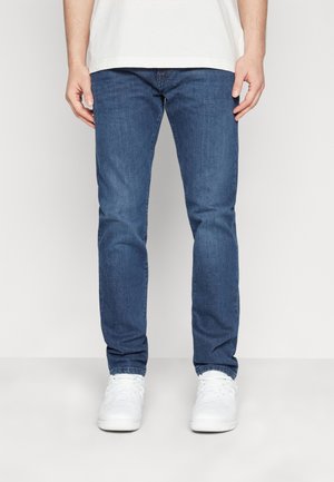 Homme portant un jean bleu, des baskets blanches et une chemise blanche, debout devant un fond clair uni.