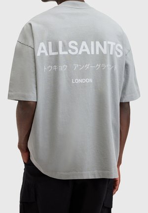 Osoba ubrana w jasnoszarą, oversize koszulkę z nadrukiem "ALLSAINTS", japońskim tekstem i "LONDON" na plecach w kolorze białym.