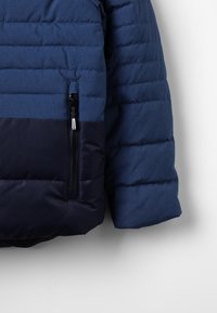 Blå puffjacka med en mörk marinblå underdel. Kvadratisk textur, sidoficka med dragkedja och slät tygfinish. Designad för värme.