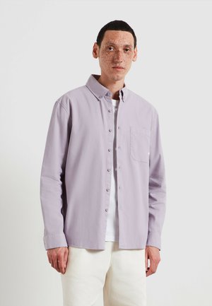 Camicia lavanda con bottoni, collo a punta, maniche lunghe e una tasca sul petto, indossata sopra una maglietta bianca e pantaloni chiari.
