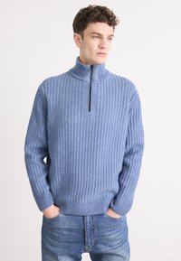 Niebieski sweter z dzianiny z kołnierzem na pół-zamek, charakteryzujący się prążkowaną teksturą i długimi rękawami. Połączony z jasnoniebieskimi dżinsami.