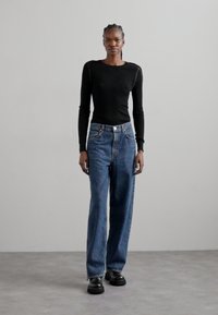 Haut noir à manches longues côtelé associé à un jean bleu loose taille haute. Le modèle porte des bottes noires chunky. Fond neutre.
