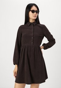 Smeđa korduroy haljina s fitted gornjim dijelom, dugim gumbima sprijeda i dugim napuhanim rukavima, s lepršavom suknjom i suptilnom teksturom u reljefu.