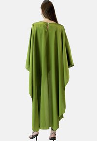 Zielony, luźny kaftan z szerokimi rękawami, z otworem w kształcie łezki z tyłu i gładkim materiałem opadającym do podłogi. Czarne szpilki widoczne poniżej.