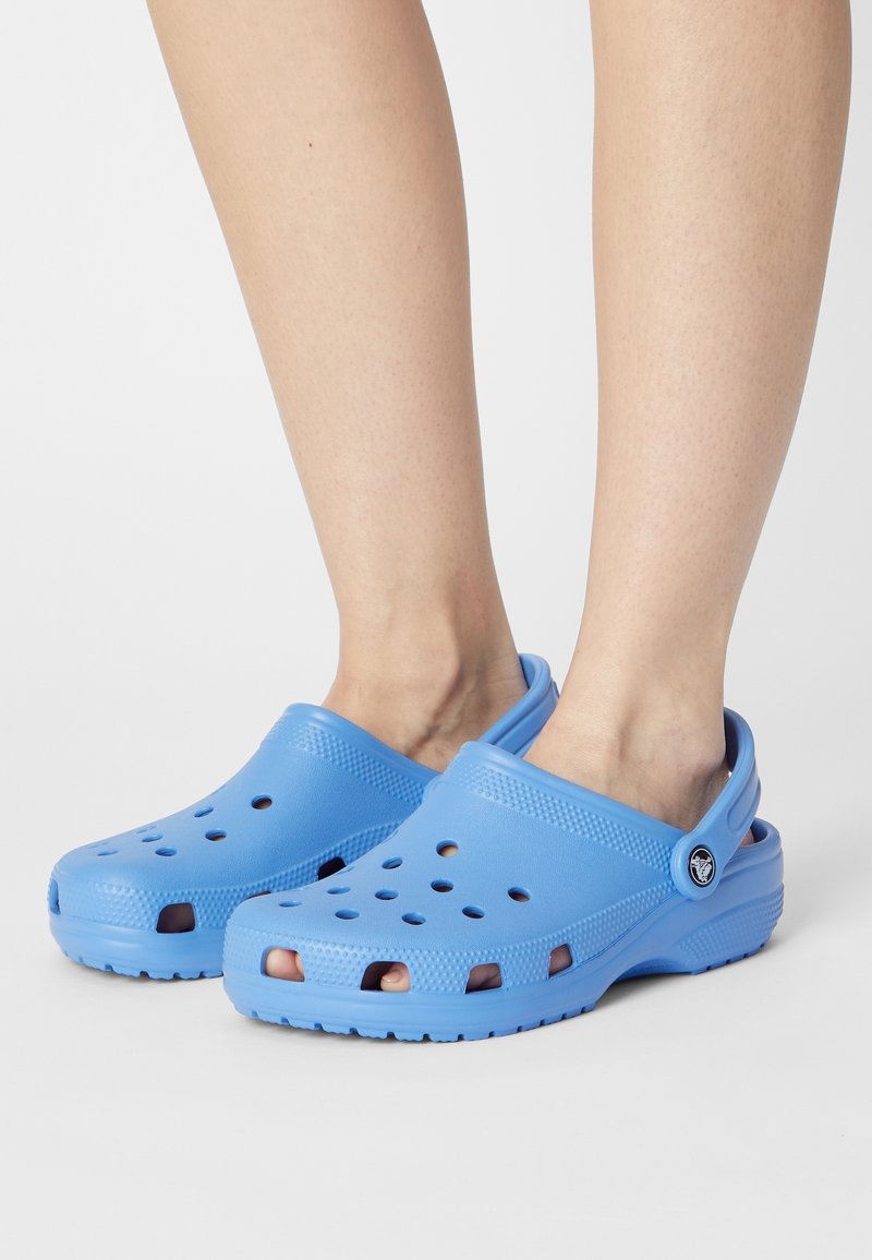 Jambes portant des sabots légers bleus clairs à enfiler, avec des trous de ventilation et des brides de talon réglables sur un fond clair uni.