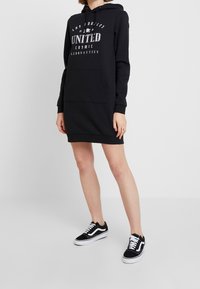 Robe hoodie noire avec poche avant, capuche et texte imprimé "Los Angeles United Cosmic Aeronautics." Associée à des baskets noir et blanc.