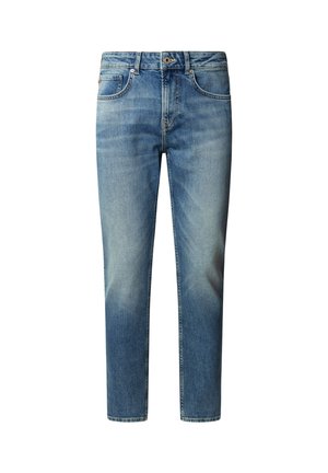 Homme portant un pull bleu marine foncé, un jean bleu clair avec une ceinture marron, et des baskets blanches, debout devant un fond clair uni.