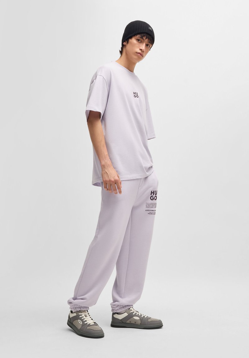 Light purple oversized t-shirt with "HUGO" logo on the chest, paired with matching joggers featuring text print and elastic cuffs, worn with sneakers.