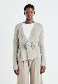 Cardigan tricotat de culoare gri deschis, cu o finisare moale și texturată, având un închidere cu fundă în față și mâneci lungi. Asortat cu o cămașă albă și pantaloni bej.