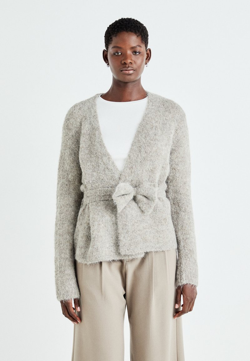 Cardigan tricotat de culoare gri deschis, cu o finisare moale și texturată, având un închidere cu fundă în față și mâneci lungi. Asortat cu o cămașă albă și pantaloni bej.