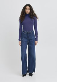 Donna con lunghi capelli ricci che indossa un dolcevita viola, jeans a vita alta e gamba larga blu e scarpe nere a punta, in piedi di fronte a uno sfondo neutro.