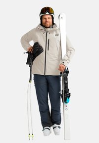 Un portascì che indossa una giacca impermeabile beige, pantaloni da sci blu scuro, guanti neri, sci bianchi e un casco con visiera amber.