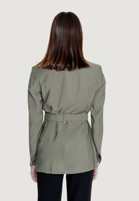 Giacca khaki con cinturino, dal taglio sartoriale e maniche lunghe, caratterizzata da una texture liscia, colletto alzato e dettagli cuciti sul retro.