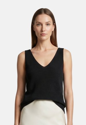 Donna con capelli castani lunghi e lisci che indossa un top nero in maglia senza maniche con scollo a V e una gonna chiara su uno sfondo semplice.