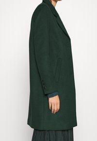 Groene wolblend jas met een op maat gemaakt ontwerp, dubbele knopen aan de voorkant, ingezette revers en zijzakken; heeft knoopmanchetten.