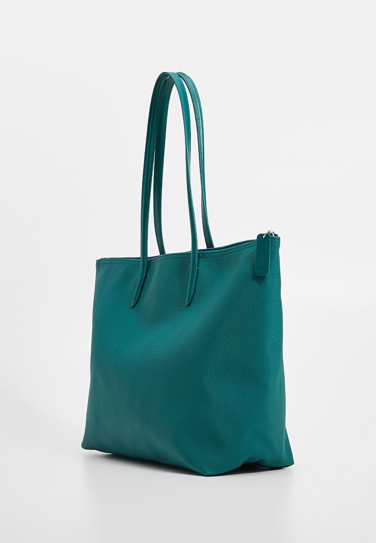 Bolso de mano de color verde azulado con una superficie texturizada, dos asas largas y un detalle de bolsillo con cremallera. Fabricado con material sintético.