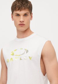 Bijela majica bez rukava s velikim, uzorkovanim logotipom "OAKLEY" u žutoj i sivoj boji. Mekana tkanina s opuštenim krojem i bočnim izrezima.