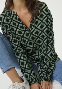 Blusa estampada en negro y verde con un diseño de diamantes, escote en V y mangas largas, combinada con jeans de color azul claro.