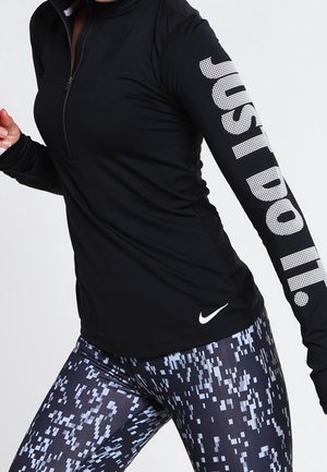 Osoba ubrana w czarną długą sportową bluzkę z napisem „JUST DO IT.” na rękawie i legginsy w cyfrowy wzór moro, w pozycji do biegu.