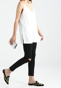 Valkoinen hihaton paita syvällä V-pääntiellä, mustat revityt skinny-farkut ja mustat ballerinat, joissa on värikkäitä kukkakirjailuja. Pitelee hopeista clutch-laukku. 