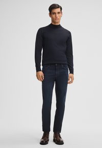 Maglione a collo alto nero a maniche lunghe abbinato a jeans slim-fit blu scuro e stivaletti stringati marroni. Design semplice, texture liscia e silhouette aderente.