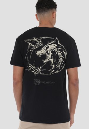Man wearing black T-shirt featuring a large graphic of a snarling wolf skull and a skeletal bird in a circular design on the back.
