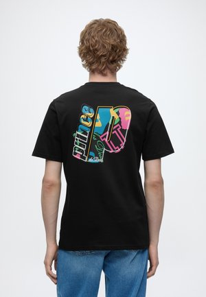 Giovane con capelli ricci che indossa una t-shirt nera con una grafica astratta colorata e testo sulla schiena, abbinata a jeans blu.