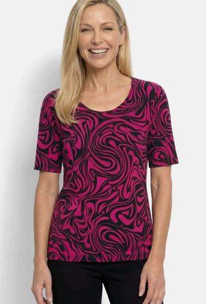 Lächelnde Frau mit glatten blonden Haaren, die ein kurzärmeliges, lila-schwarz abstrakt gemustertes Oberteil und schwarze Hose trägt.