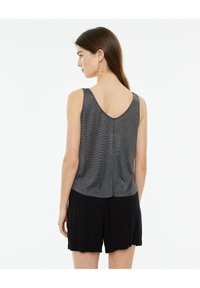 Top gris sin mangas con escote redondeado y tejido texturizado. La modelo lo lleva con pantalones cortos negros, vista desde atrás.