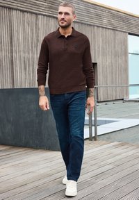 Homme avec des tatouages marchant en extérieur portant un polo marron à manches longues, un jean bleu et des baskets blanches sur une terrasse en bois près d'un bâtiment moderne.