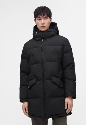 Jeune homme portant un long manteau d'hiver noir matelassé avec fermeture éclair sur le devant, col montant, capuche et poches à rabat sur un fond blanc uni.