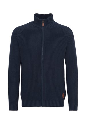 Mørkeblå, ribbestrikket genser med høy krage og glidelås foran. Har en brun lærlogoaccent nederst til venstre.