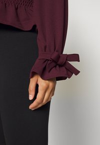 Burgunderfarbene Bluse mit gerafftem Ärmel-Detail und Schleifenverschluss am Bund, kombiniert mit enganliegenden schwarzen Leggings. Glatte Stofftextur.