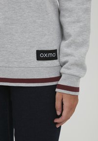 Sweat à capuche gris avec un ourlet et des poignets côtelés bordeaux rayés, arborant une étiquette noire avec le logo "oxmo" fixé au bas.