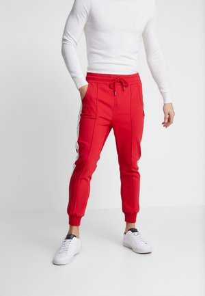 Mann in roten Jogginghosen mit seitlichen Streifen, weißen Sneakers und einem enganliegenden weißen Langarmoberteil, stehend vor einem weißen Hintergrund.