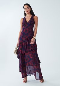 Maxi dress in sheer fabric featuring a deep V neckline, layered ruffles, and a floral pattern in deep red and navy. Includes strappy sandals.