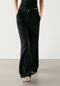 Zwarte, wijde broek gemaakt van een zachte stof, met knoopsluiting en zijzakken. Gemaakt in combinatie met een aansluitend zwart topje en sandalen.