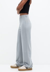 Personne portant un pantalon large taille haute bleu clair, un haut noir sans manches, et des baskets blanches à rayures bleues, debout de profil les bras croisés.