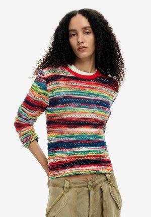 Kobieta o kręconych włosach, ubrana w wielokolorowy sweter w paski i beżowe spodnie, stojąca z jedną ręką za plecami.