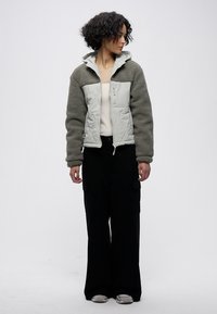 Světlé zelená a šedá fleecová bunda se zipem vpředu, texturovaný materiál, přiléhavý design a dlouhé rukávy, nošená přes krémový top a černé kalhoty.