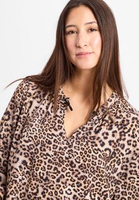 Frau mit langen, glatten braunen Haaren, die ein V-Ausschnitt-Oberteil im Leopardenprint trägt, mit neutralem Gesichtsausdruck vor einem weißen Hintergrund.