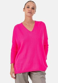 Pull à col en V rose vif avec poignets et ourlet côtelés, présentant une coupe ample et une texture douce, accompagné d'un pantalon gris clair.