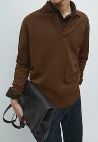 Brauner Strickpullover mit V-Ausschnitt und Kragen, kombiniert mit einem dunklen Hemd. Schwarze Ledertasche unter einem Arm gehalten. Dunkle Jeans aus Denim.